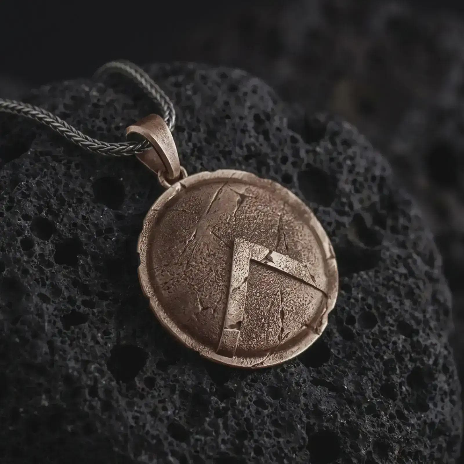 Bronze Spartan shield pendant necklace on textured volcanic rock, close-up studio shot highlighting the cracked round shield, raised lambda symbol, battle-worn texture, and antique silver contrast.