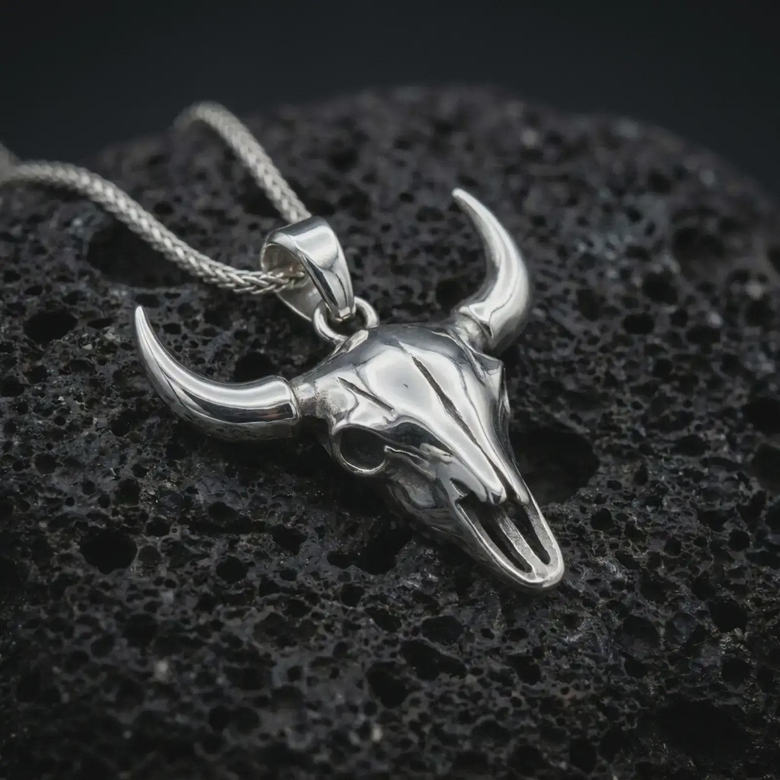 Polished silver bull skull pendant necklace on textured volcanic rock, centered macro jewelry photo with detailed horn structure, skull texture, and bright silver highlights.