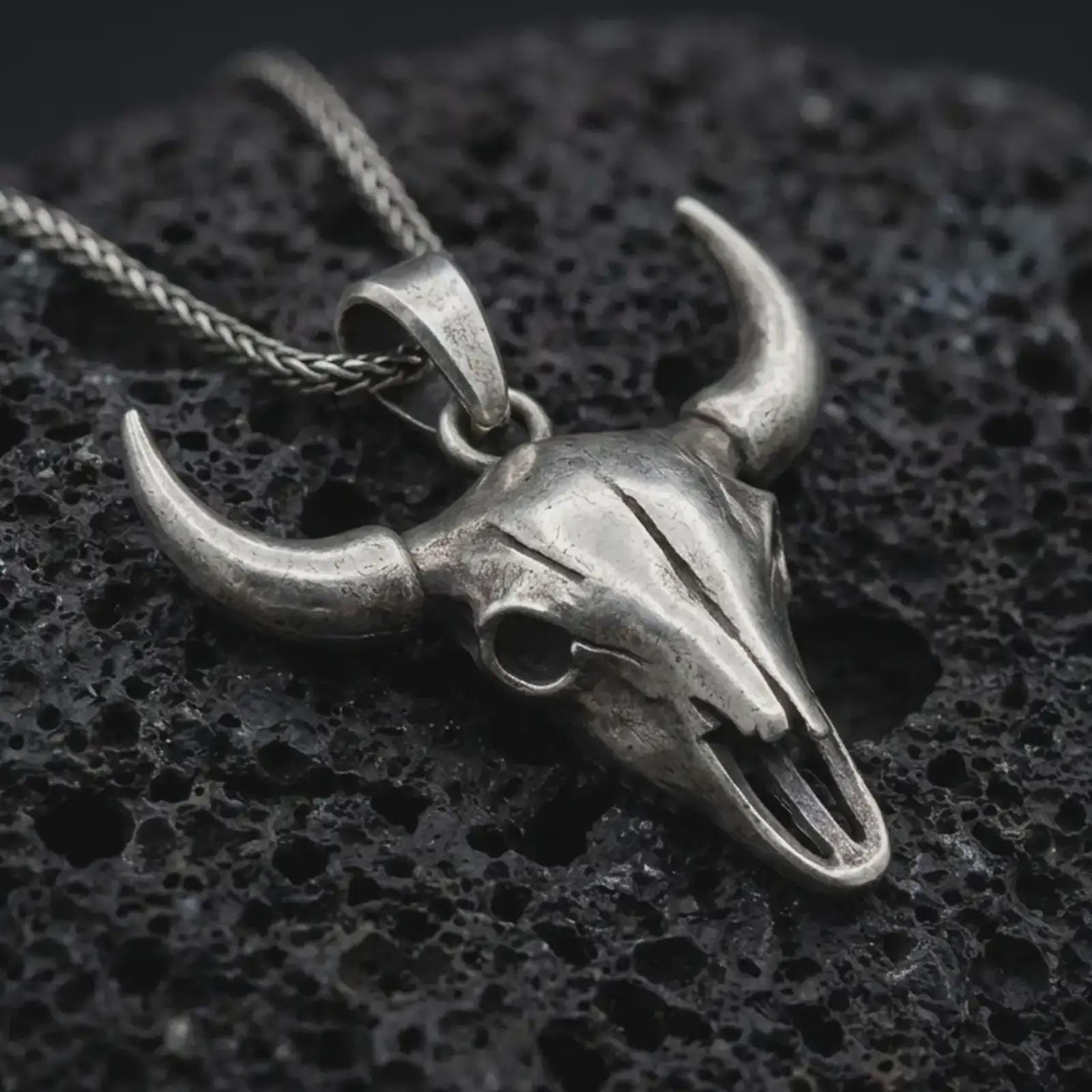Oxidized bull skull pendant necklace on textured volcanic rock, close-up studio shot highlighting the long horns, hollow skull form, western design, and antique silver contrast.
