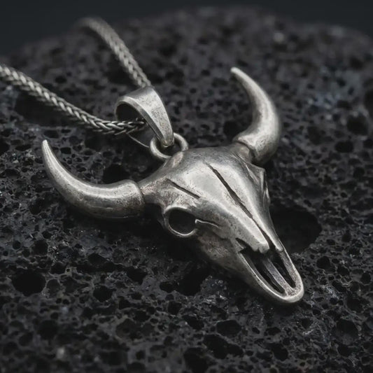 Oxidized bull skull pendant necklace on textured volcanic rock, close-up studio shot highlighting the long horns, hollow skull form, western design, and antique silver contrast.