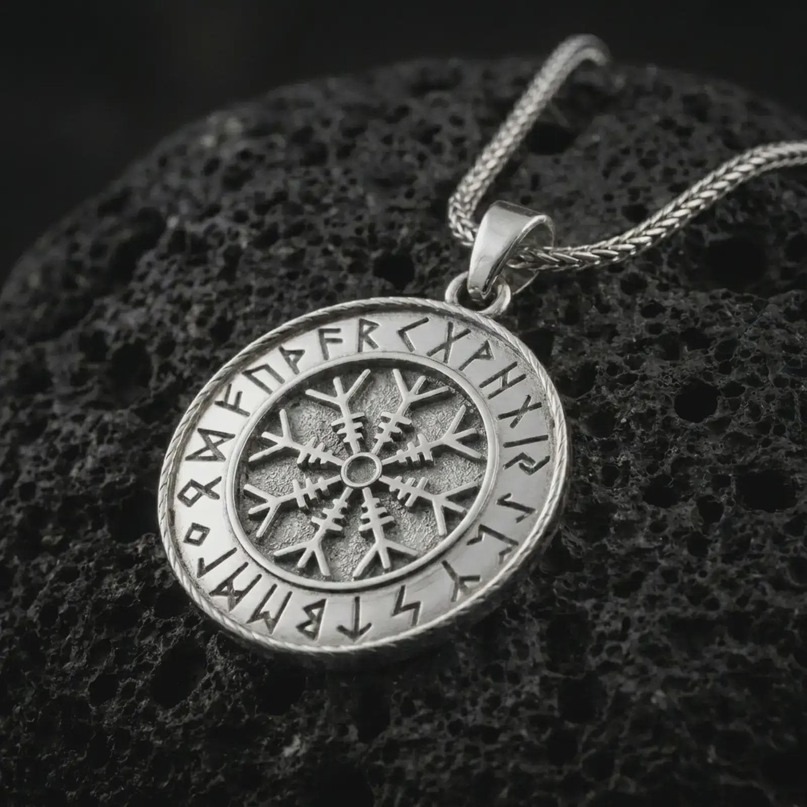 Polished silver Vegvisir Viking rune pendant necklace on textured volcanic rock, centered macro jewelry photo with detailed compass relief and bright silver highlights.
