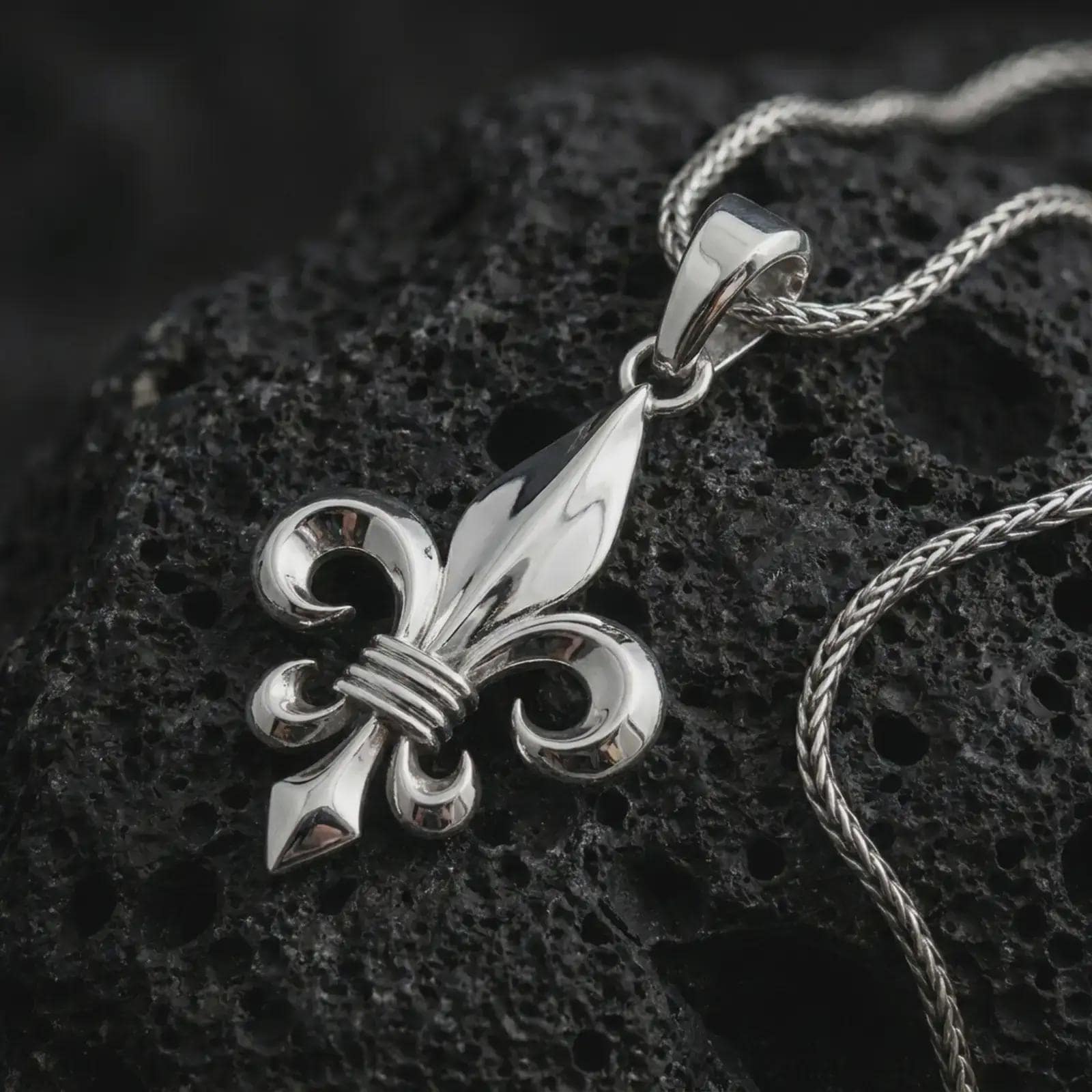Polished silver fleur-de-lis pendant necklace on textured volcanic rock, centered macro jewelry photo with detailed French Gothic form and bright silver highlights.