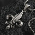 Load image into Gallery viewer, Oxidized fleur-de-lis pendant necklace on textured volcanic rock, close-up studio shot highlighting the royal lily shape, curled heraldic arms, sculpted center blade, and antique silver contrast.

