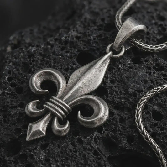 Oxidized fleur-de-lis pendant necklace on textured volcanic rock, close-up studio shot highlighting the royal lily shape, curled heraldic arms, sculpted center blade, and antique silver contrast.