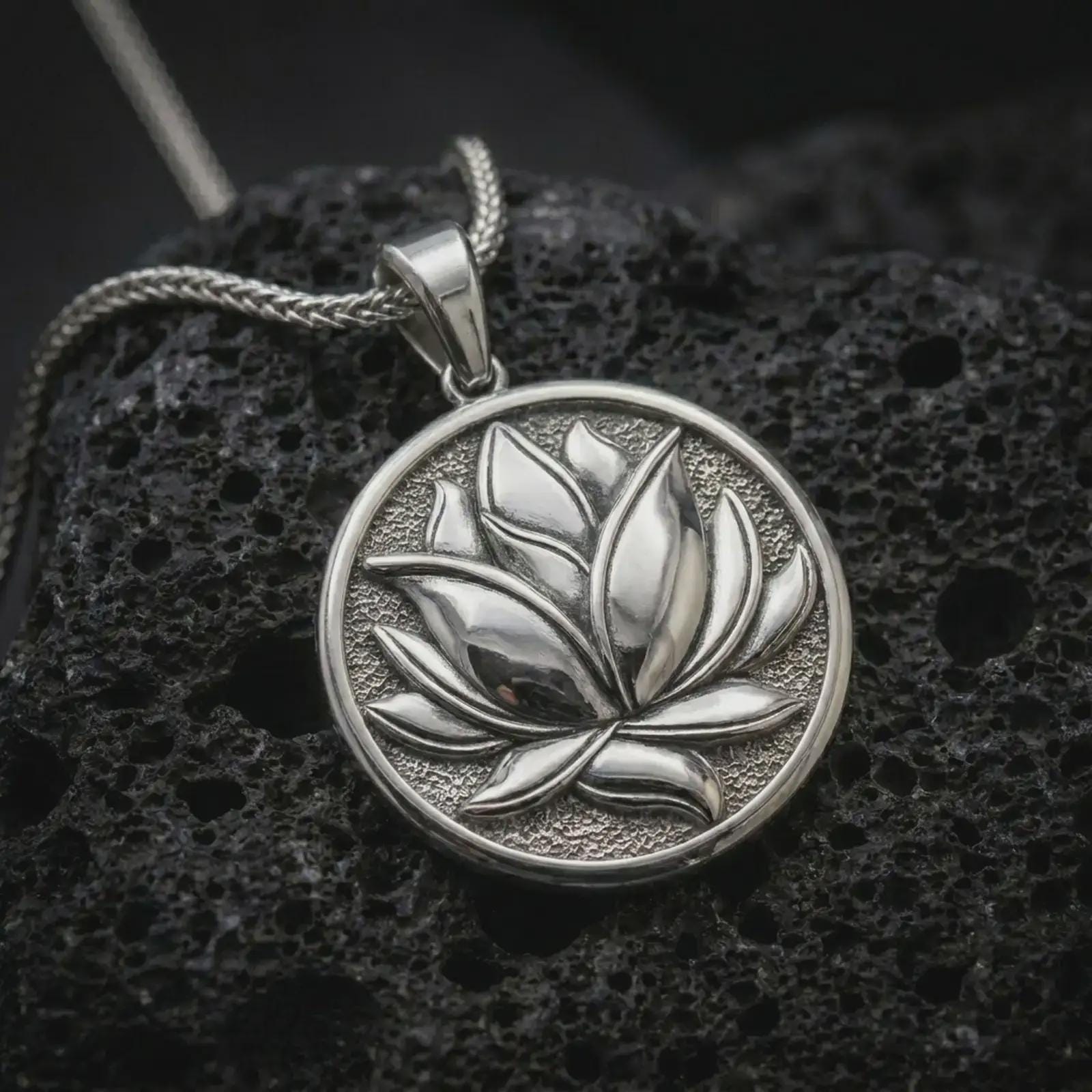 Polished silver lotus flower pendant necklace on textured volcanic rock, centered macro jewelry photo with detailed petal relief and bright silver highlights.
