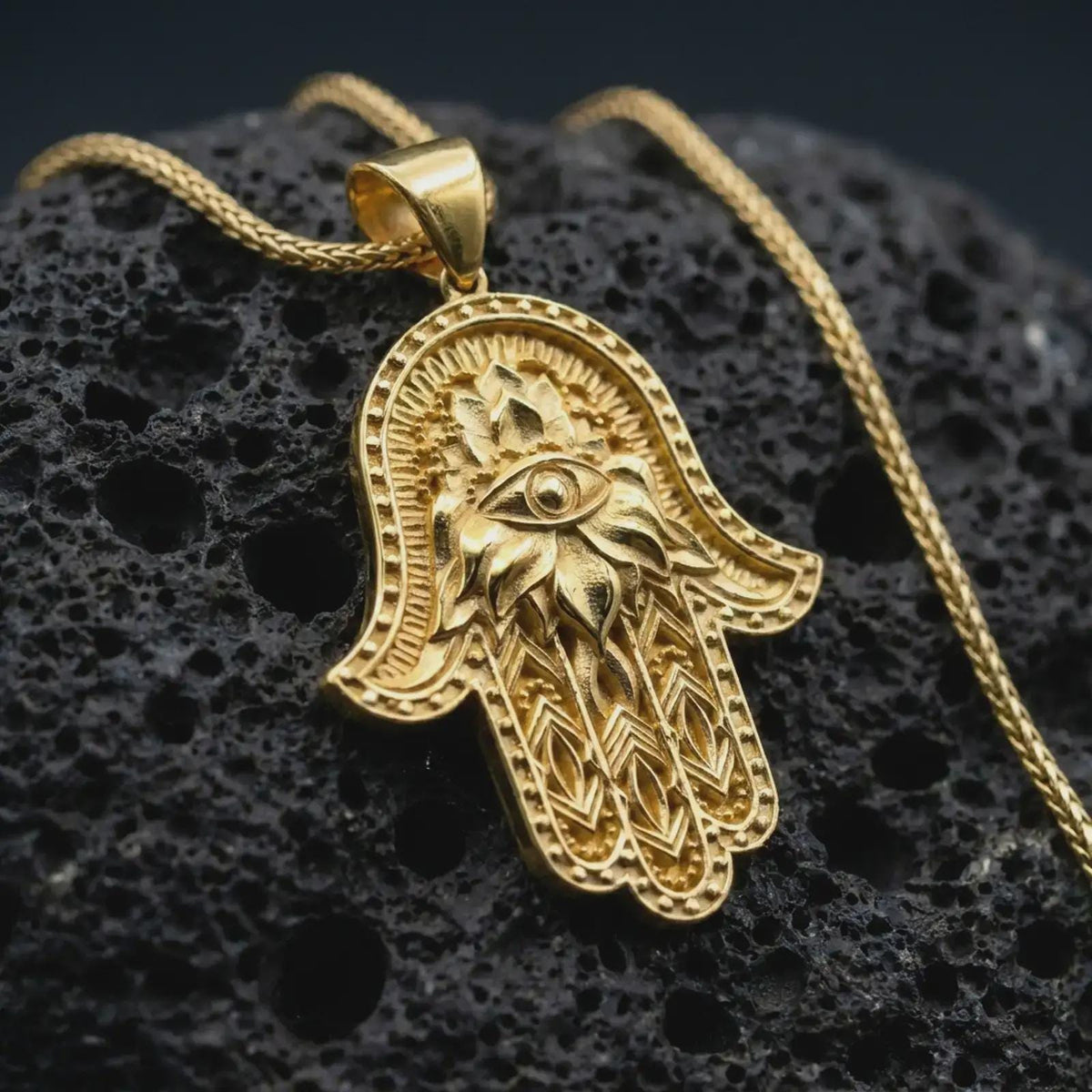 Gold plated hamsa hand pendant necklace on textured volcanic rock, premium close-up studio composition emphasizing the protective hand symbol, lotus details, and spiritual amulet design.