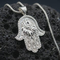 Load image into Gallery viewer, Polished silver hamsa hand pendant necklace on textured volcanic rock, centered macro jewelry photo with detailed evil eye charm, floral pattern, and bright silver highlights.
