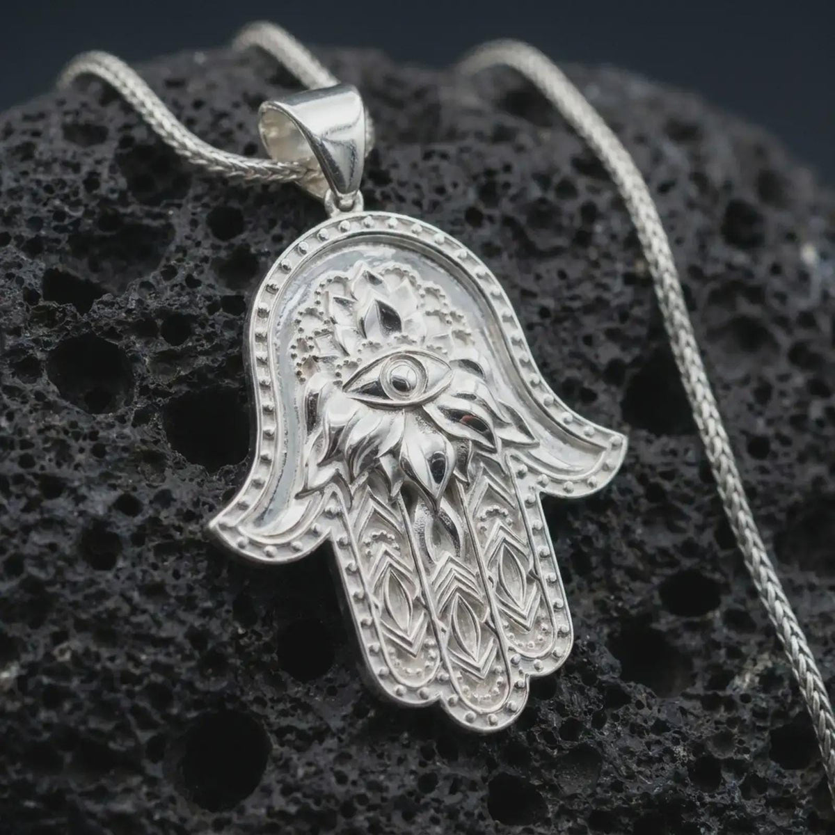 Polished silver hamsa hand pendant necklace on textured volcanic rock, centered macro jewelry photo with detailed evil eye charm, floral pattern, and bright silver highlights.