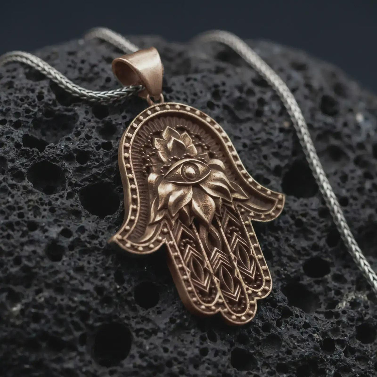 Bronze hamsa hand pendant necklace on textured volcanic rock, close-up studio shot highlighting the evil eye center, lotus flower relief, ornate hand shape, and antique silver contrast.