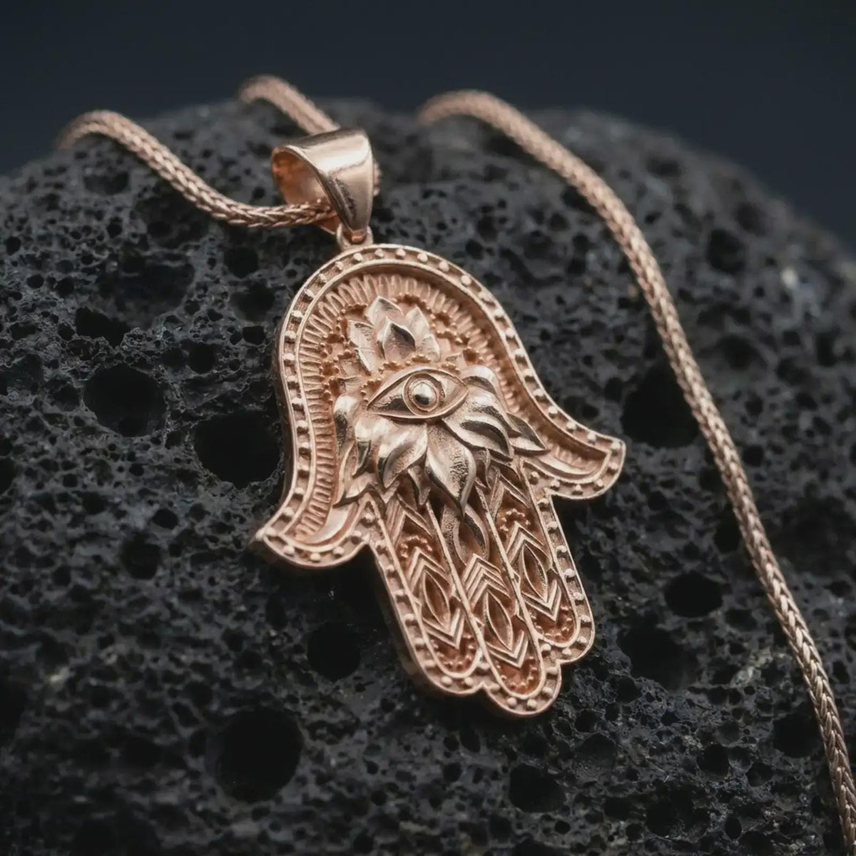 Rose gold plated hamsa hand pendant necklace on textured volcanic rock, detailed macro product shot with soft warm reflections and sacred evil eye symbolism.