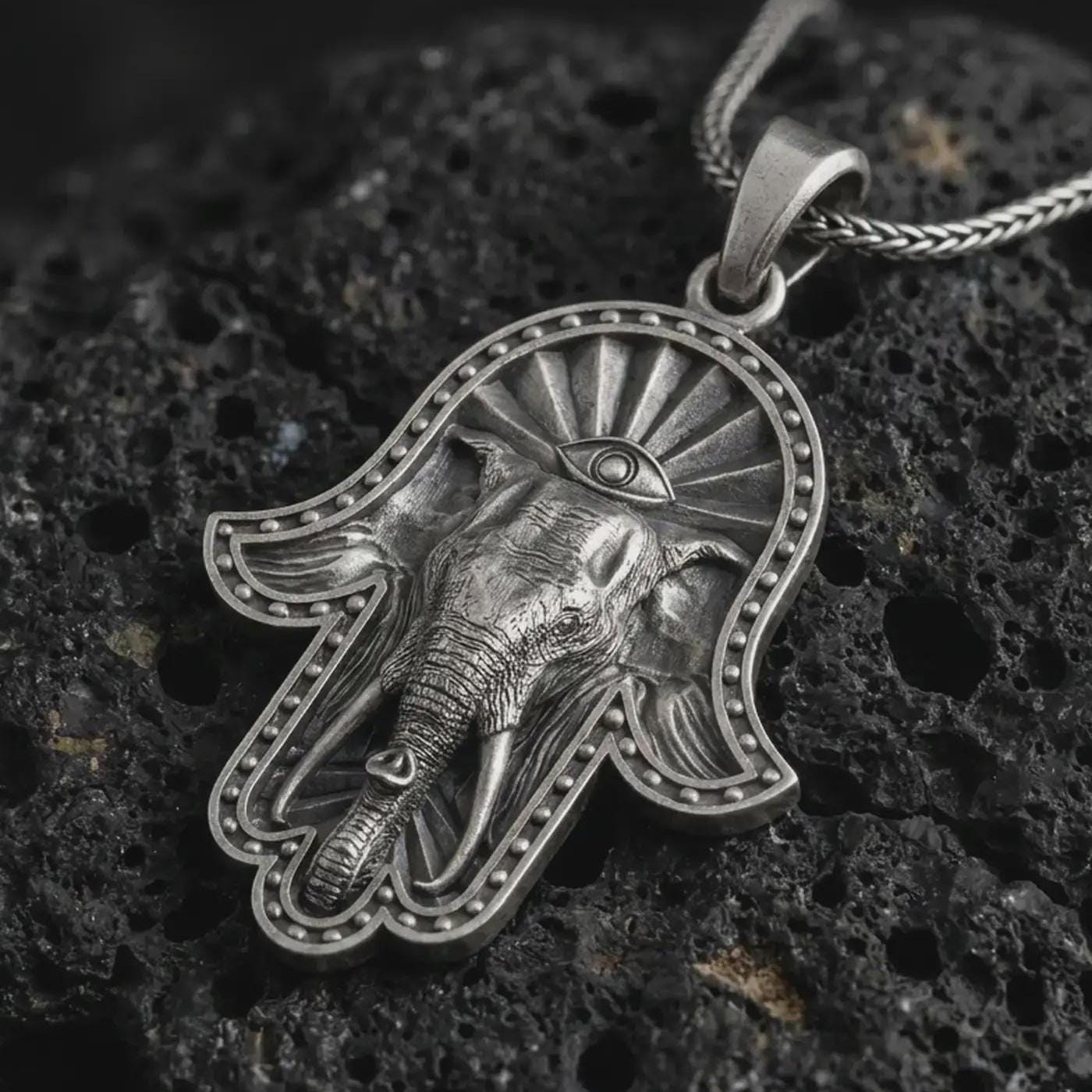 Oxidized hamsa elephant pendant necklace on textured volcanic rock, close-up studio shot highlighting the elephant head relief, third eye symbol, beaded hand frame, and antique silver contrast.