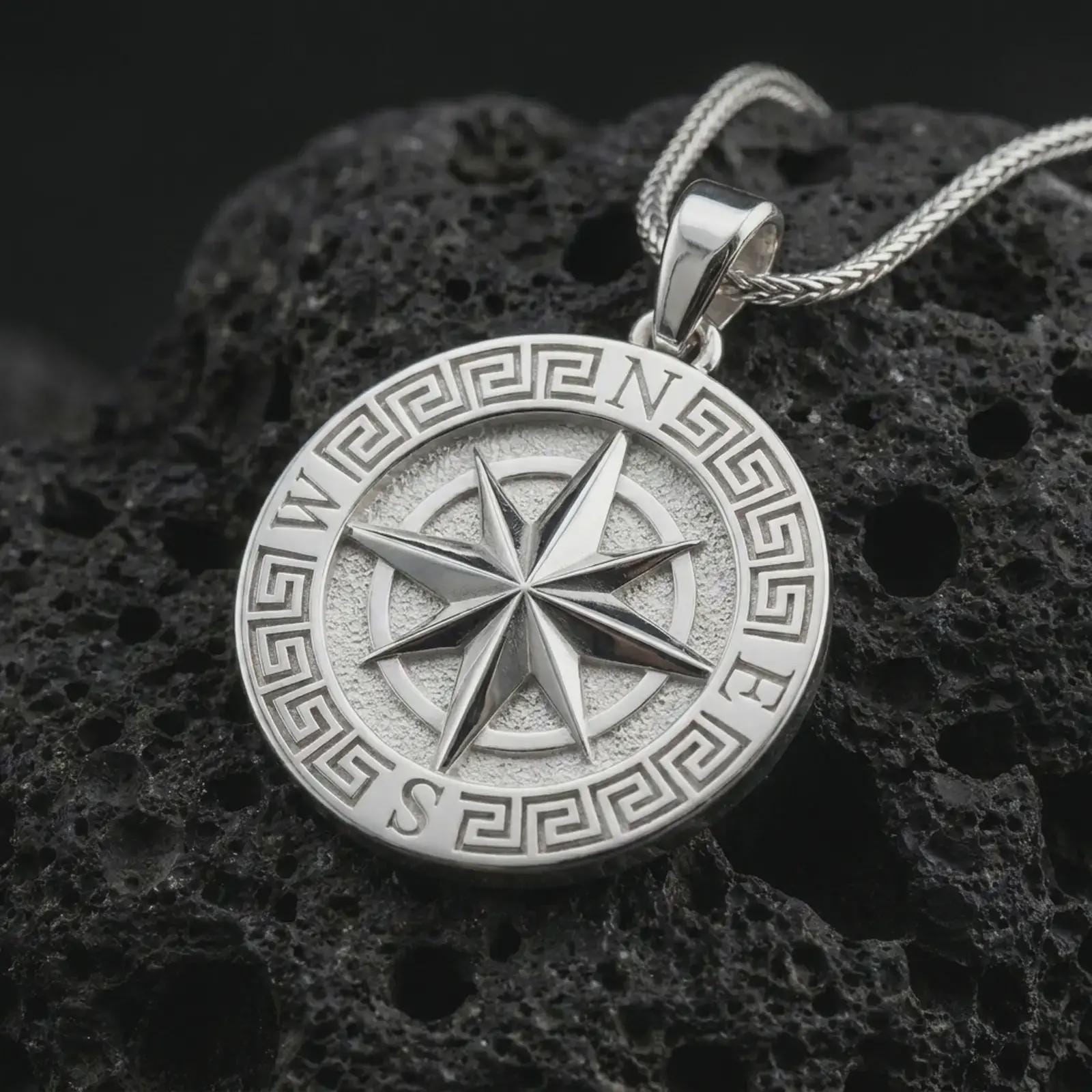 Polished silver Greek compass pendant necklace on textured volcanic rock, centered macro jewelry photo with detailed directional star relief and bright silver highlights.