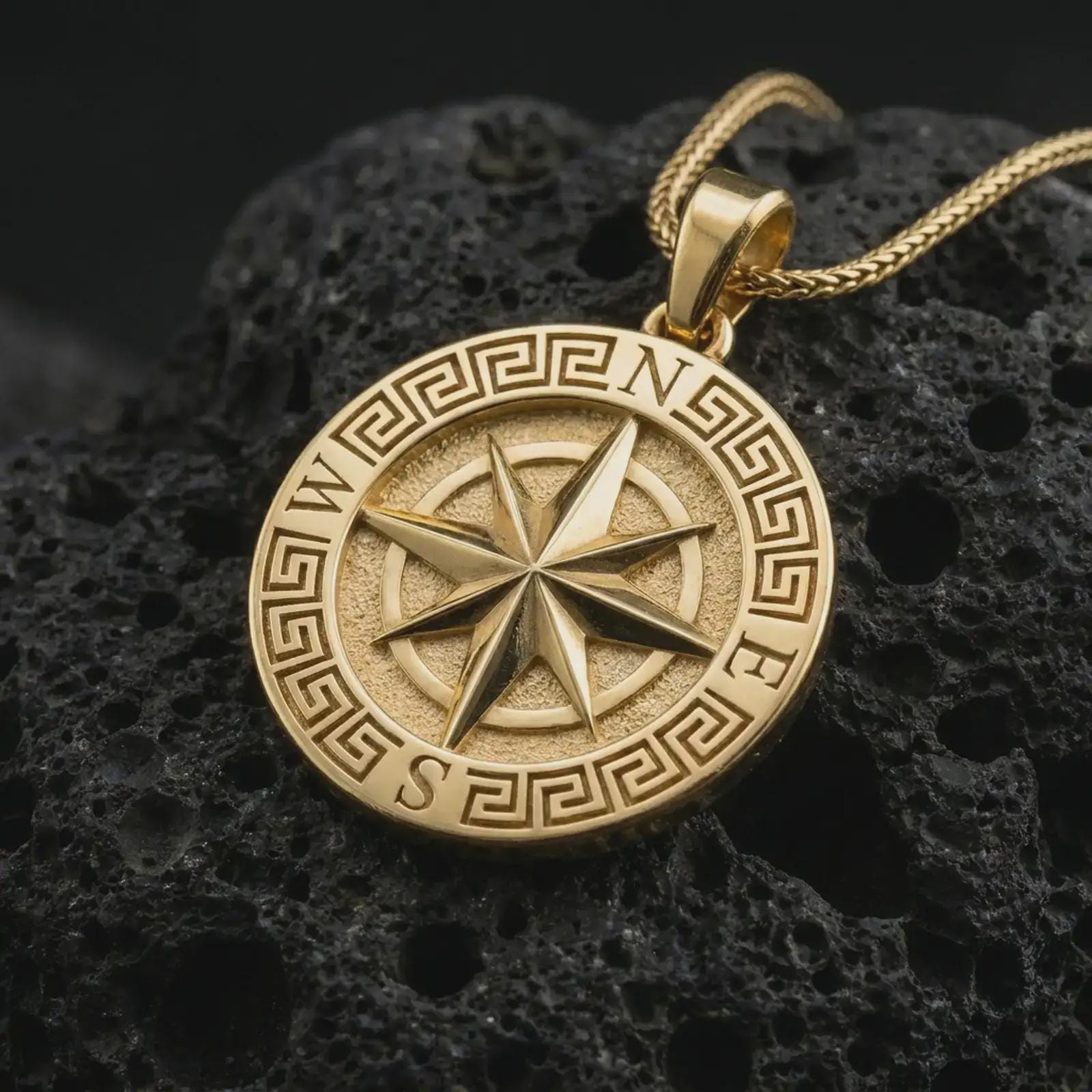 Gold plated Greek compass pendant necklace on textured volcanic rock, premium close-up studio composition emphasizing the navigation symbol, Greek meander border, and graduation gift design.