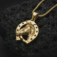 Load image into Gallery viewer, Gold plated horseshoe pendant necklace on textured volcanic rock, premium close-up studio composition emphasizing the good luck symbol, horse head detail, and protective charm design.
