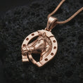 Load image into Gallery viewer, Rose gold plated horseshoe pendant necklace on textured volcanic rock, detailed macro product shot with soft warm reflections and symbolic fortune-inspired styling.
