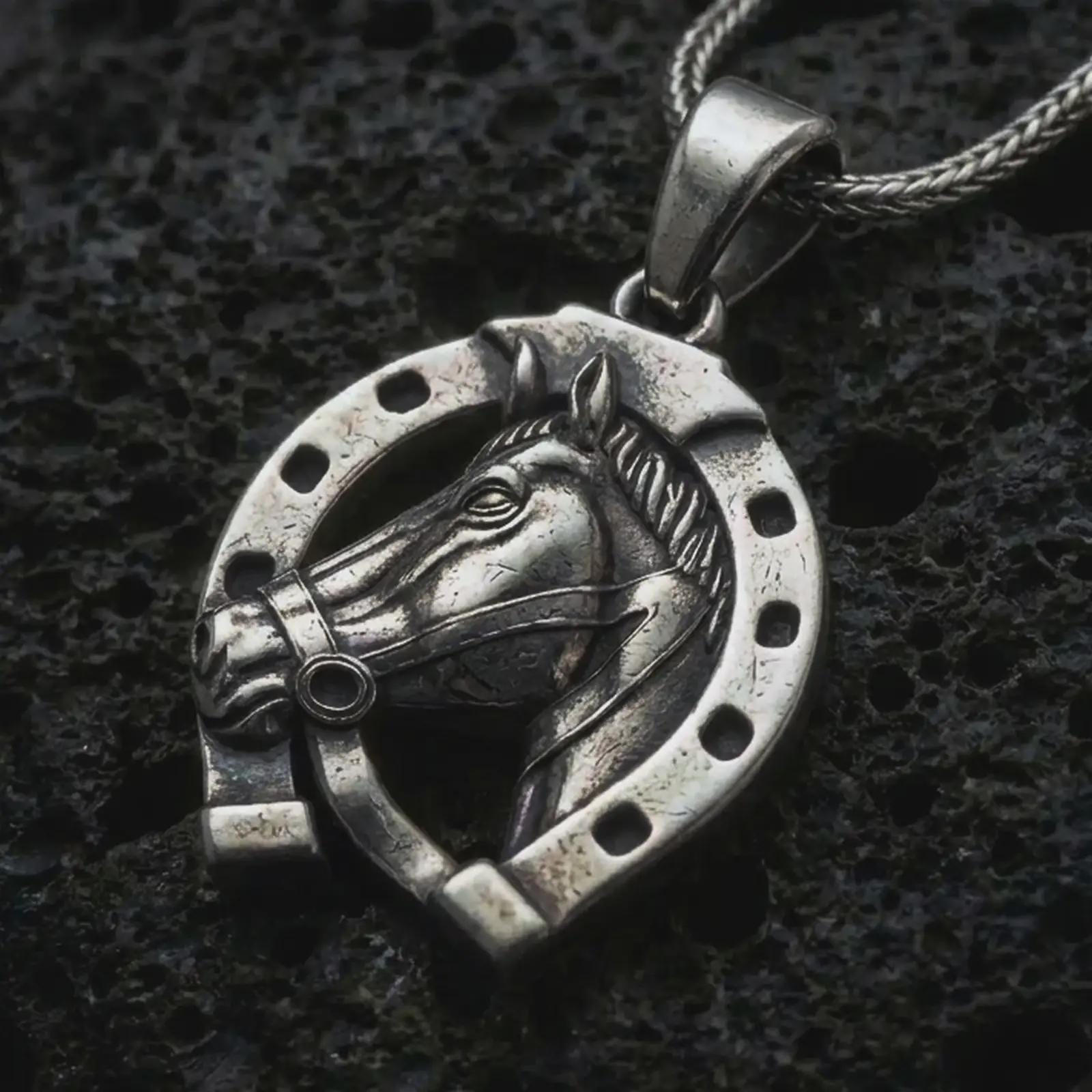Oxidized horseshoe pendant necklace on textured volcanic rock, close-up studio shot highlighting the horse head relief, lucky horseshoe frame, openwork design, and antique silver contrast.