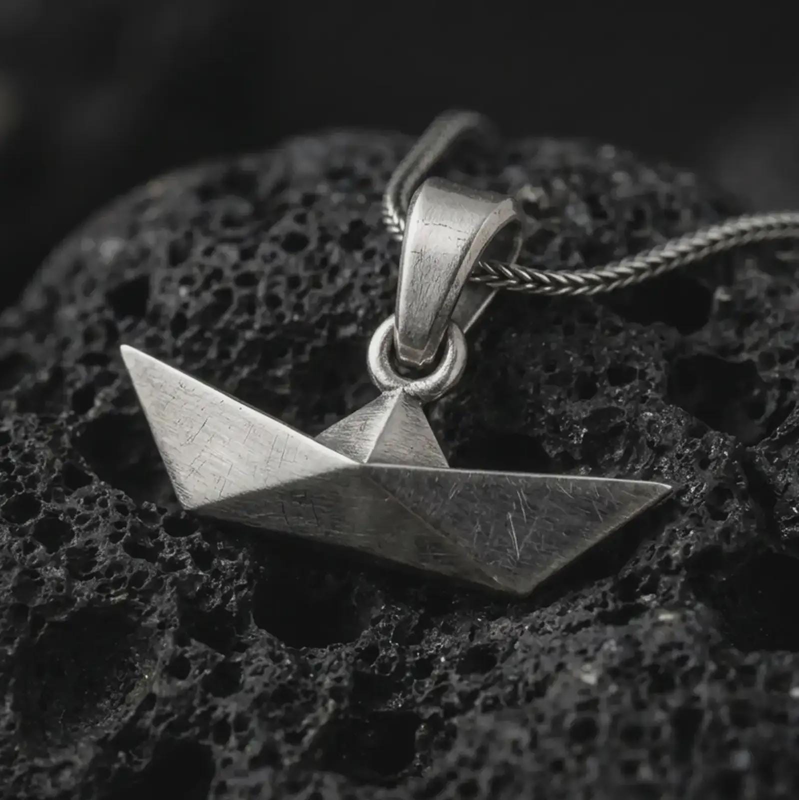 Oxidized origami boat pendant necklace on textured volcanic rock, close-up studio shot highlighting the folded paper boat shape, geometric angles, brushed surface, and antique silver contrast.