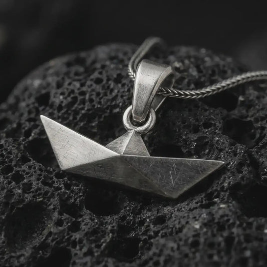 Oxidized origami boat pendant necklace on textured volcanic rock, close-up studio shot highlighting the folded paper boat shape, geometric angles, brushed surface, and antique silver contrast.