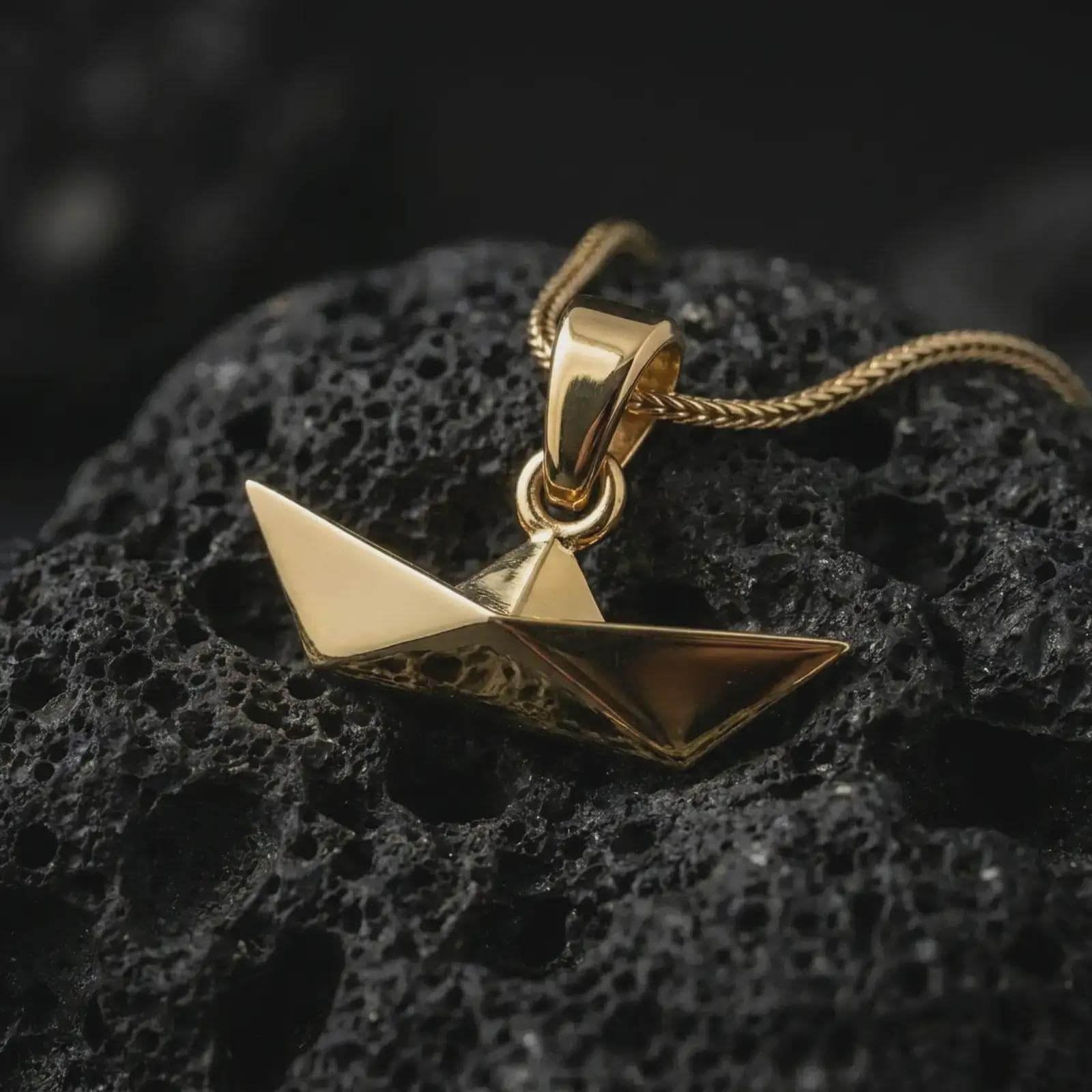Gold plated origami boat pendant necklace on textured volcanic rock, premium close-up studio composition emphasizing the minimalist paper boat design, sharp geometry, and symbolic journey charm.