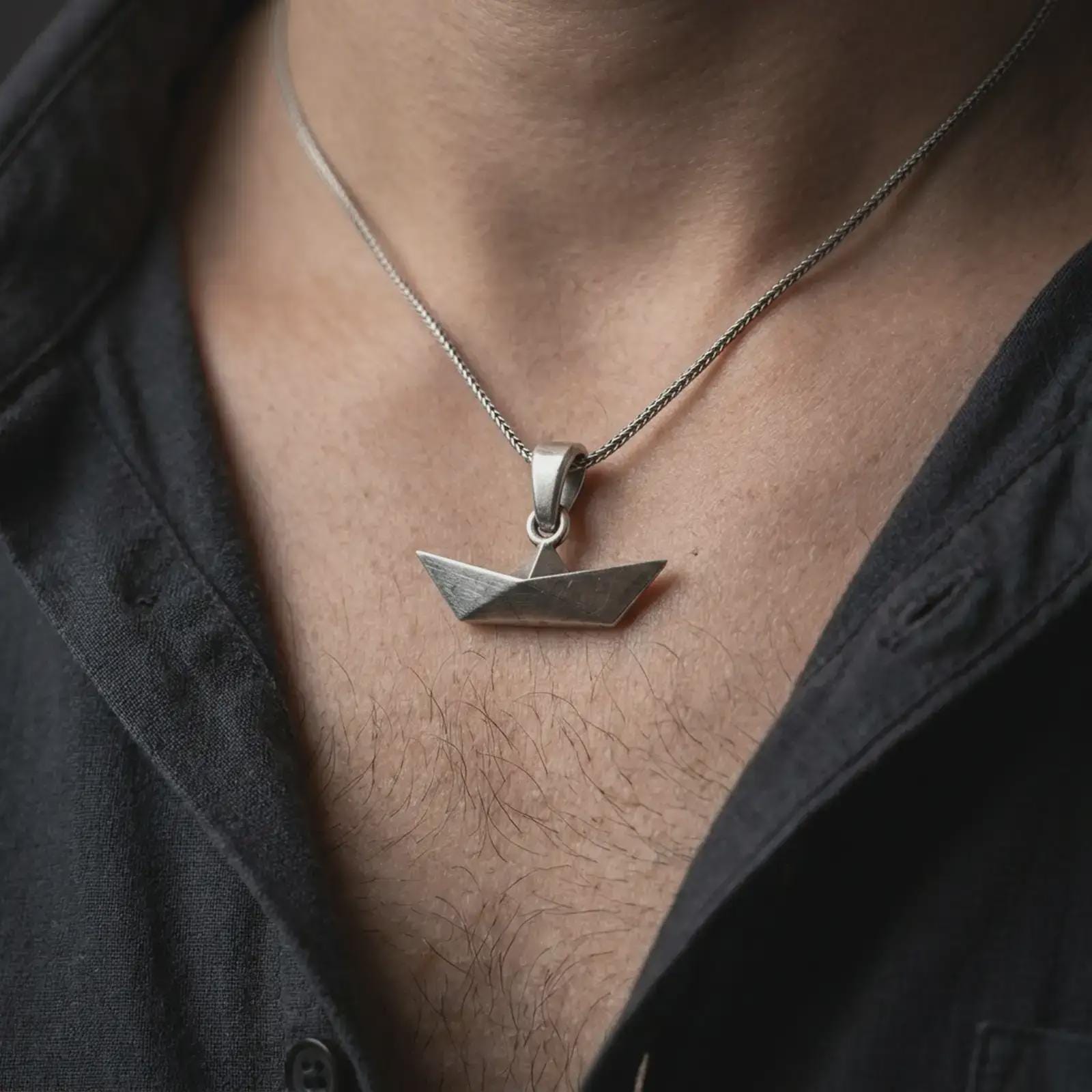 Oxidized origami boat pendant necklace worn on the chest, close-up lifestyle jewelry photo showing the geometric boat charm on a spiga chain with dark clothing.