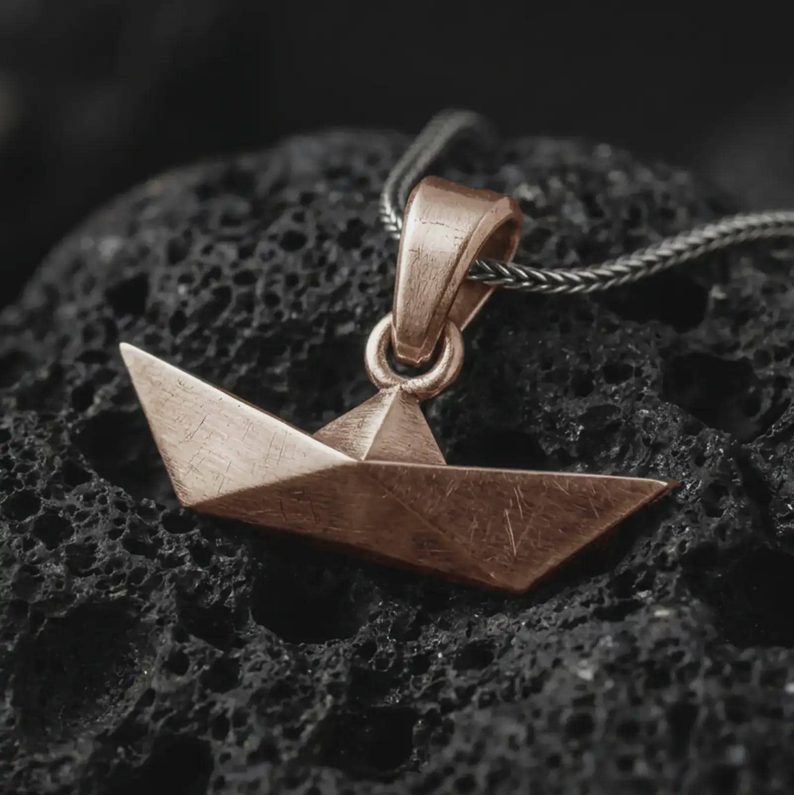 Bronze origami boat pendant necklace on textured volcanic rock, close-up studio shot highlighting the folded paper boat shape, geometric angles, brushed surface, and antique silver contrast.