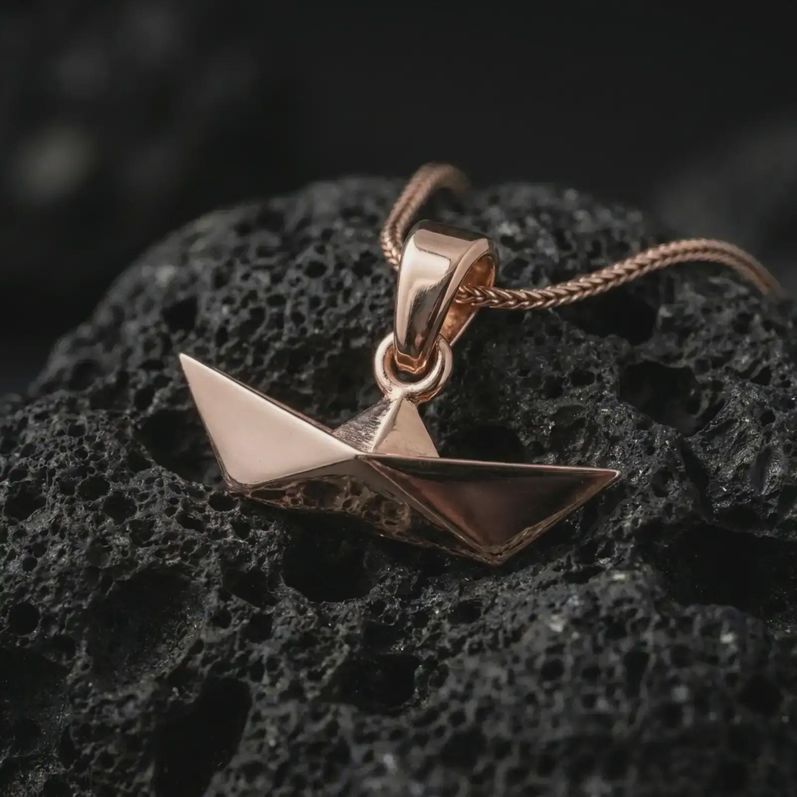 Rose gold plated origami boat pendant necklace on textured volcanic rock, detailed macro product shot with soft warm reflections and elegant folded boat styling.