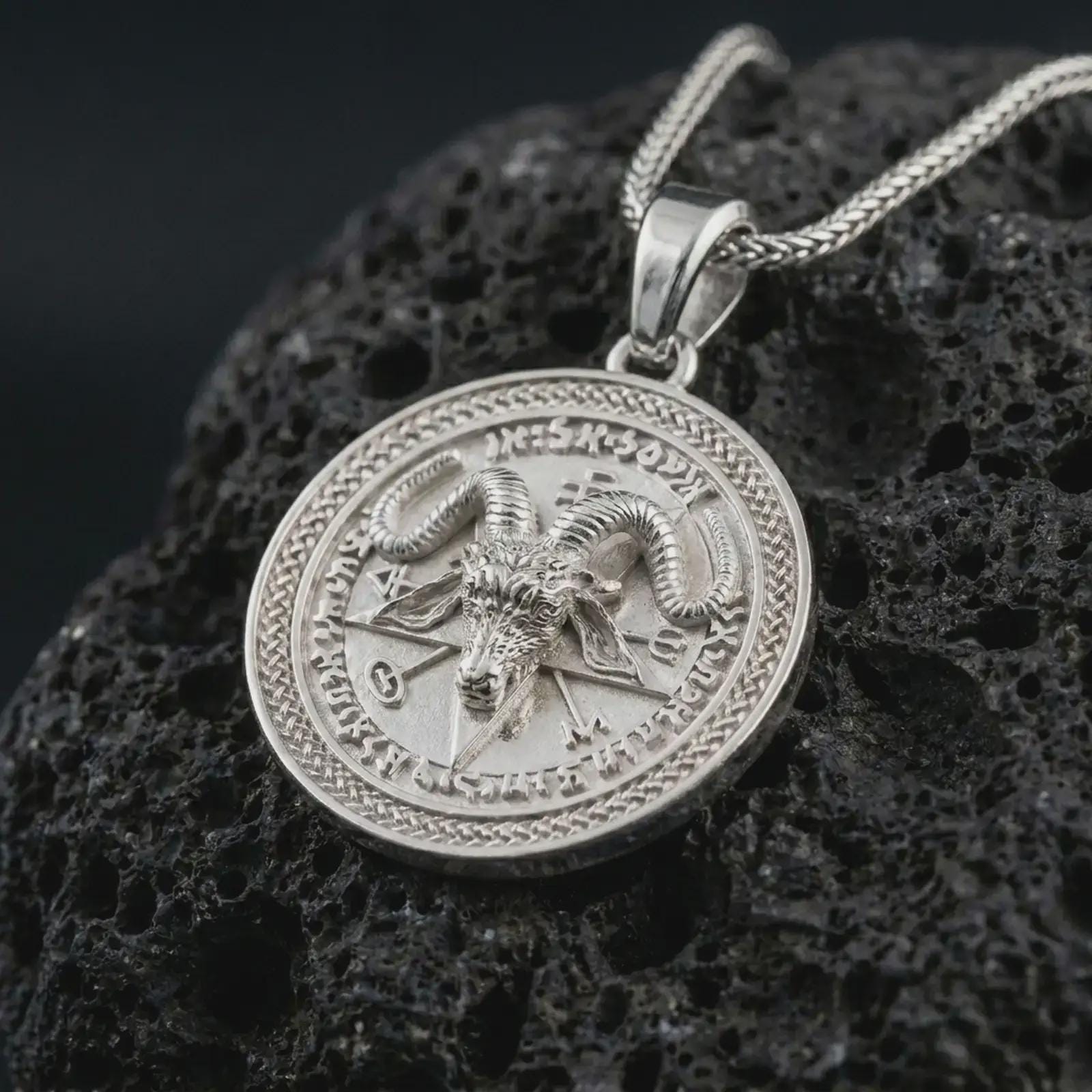 Polished silver Baphomet pendant necklace on textured volcanic rock, centered macro jewelry photo with detailed ram horns, symbolic lettering, and bright silver highlights.