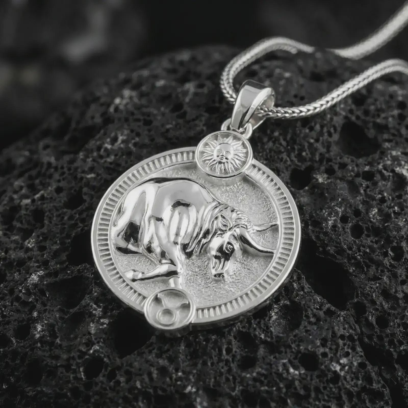 Polished silver Taurus zodiac pendant necklace on textured volcanic rock, centered macro jewelry photo with detailed bull horoscope relief and bright silver highlights.
