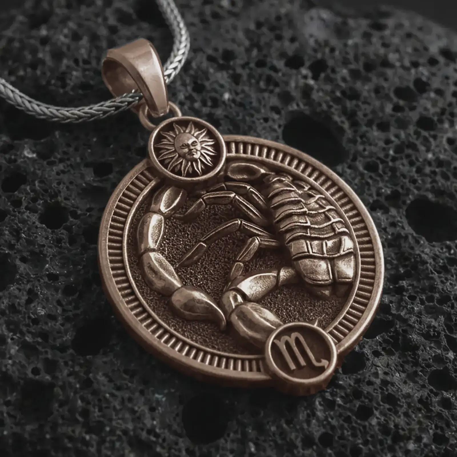 Bronze Scorpio zodiac pendant necklace on textured volcanic rock, close-up studio shot highlighting the scorpion relief, sun detail, Scorpio symbol, and antique silver contrast.