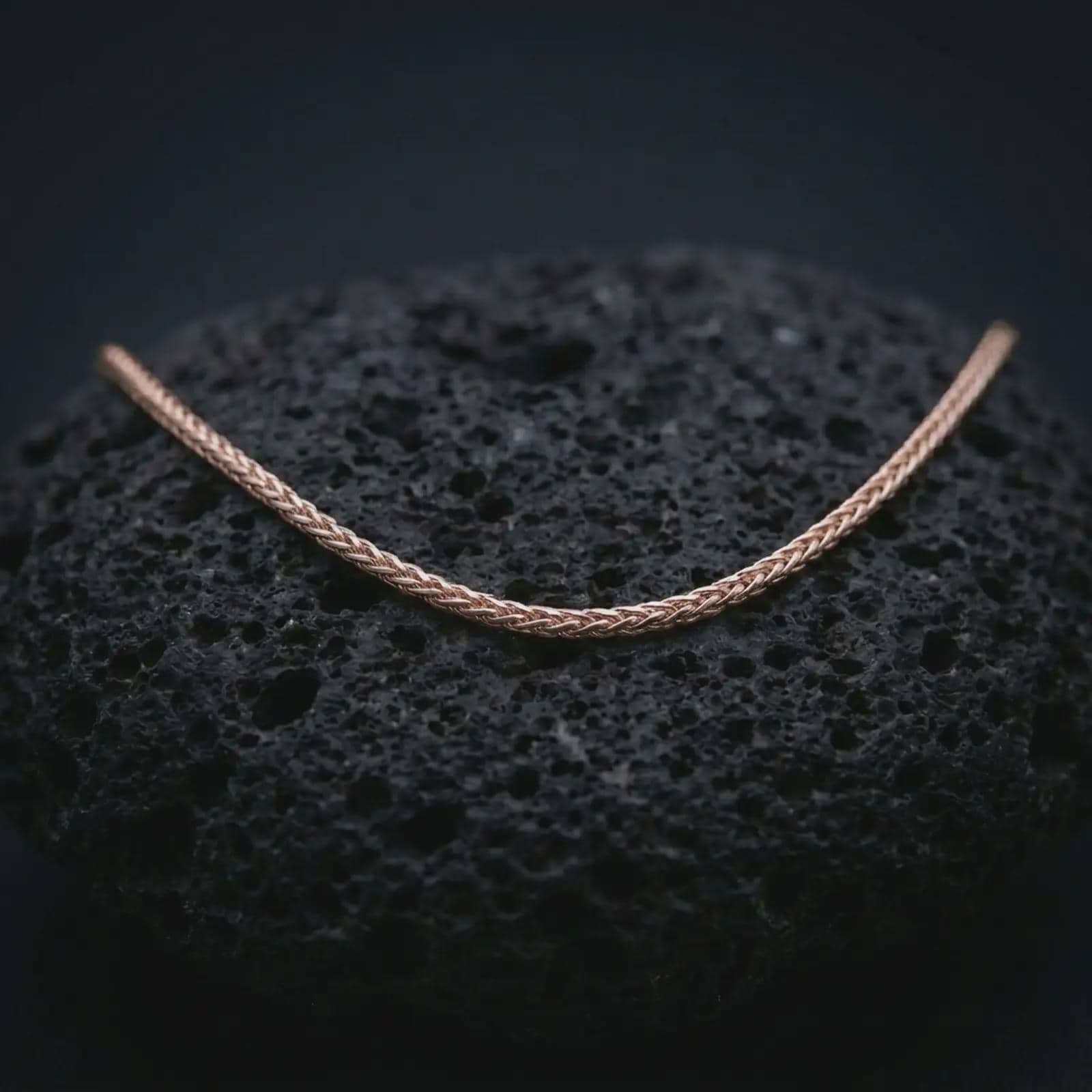 A rose gold plated foxtail chain necklace is photographed on porous volcanic rock in a close-up studio composition with refined luxury lighting and smooth chain texture.