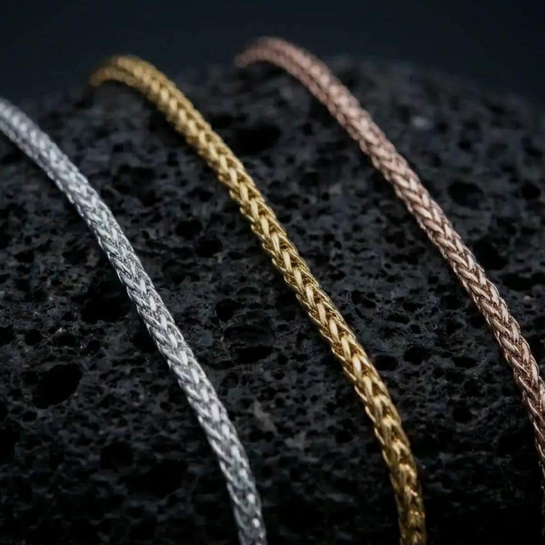 A polished silver, gold plated, and rose gold plated foxtail chain necklace rests on dark volcanic rock in a macro studio close-up, showing the detailed woven silver texture and premium finish.