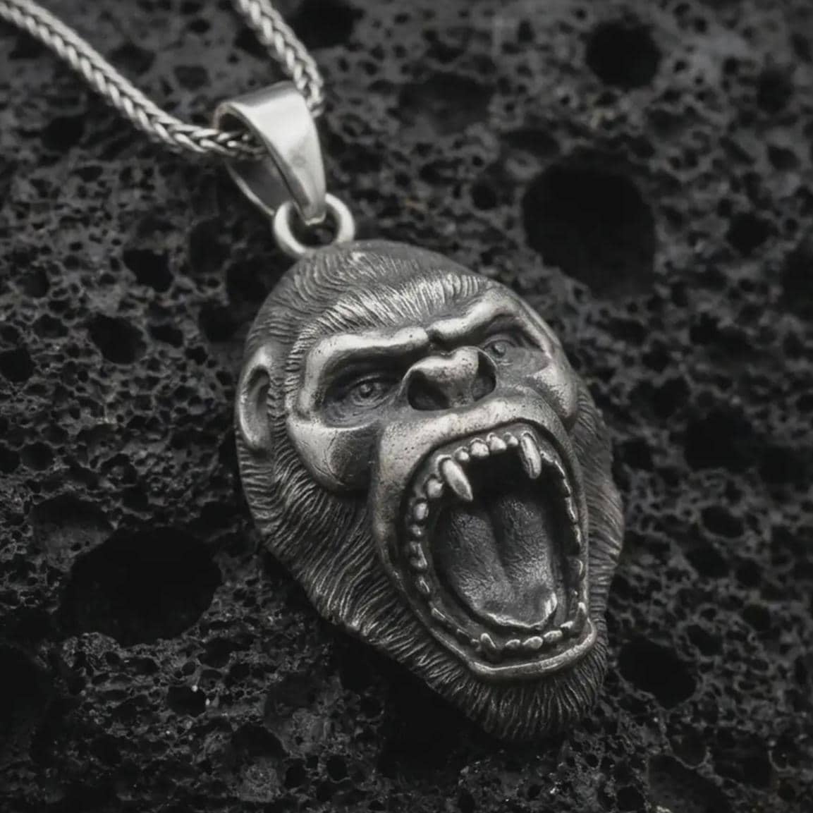 An oxidized gorilla pendant rests on dark volcanic rock in a macro studio close-up, showing a fierce roaring ape head with detailed teeth and fur texture.