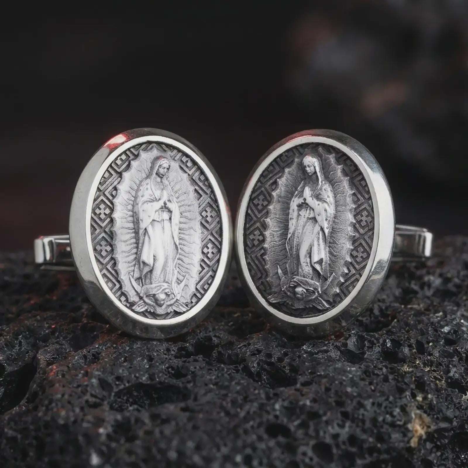 An oxidized Our Lady of Guadalupe cufflink pair rests on dark volcanic rock in a macro studio close-up, showing the Virgin Mary relief in oval frames.