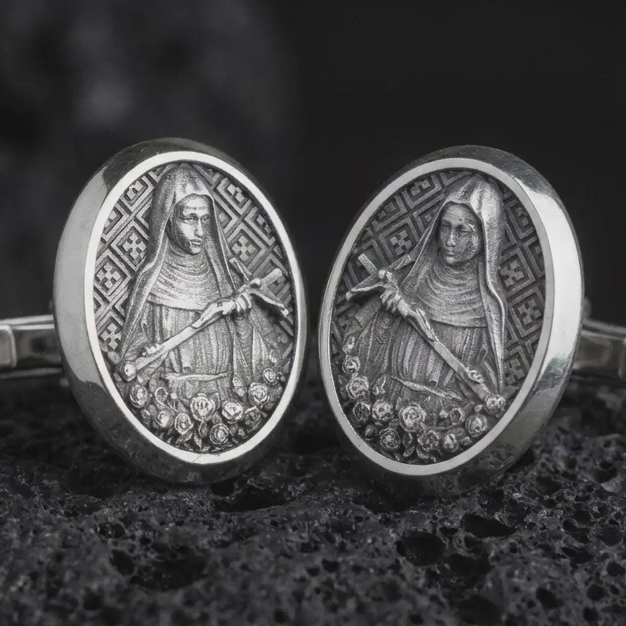 An oxidized Mary Magdalene cufflink pair rests on dark volcanic rock in a macro studio close-up, showing a detailed saint relief inside oval frames.