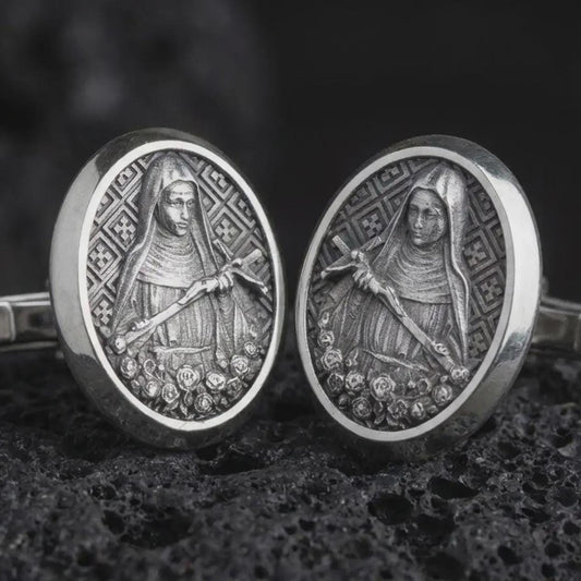 An oxidized Mary Magdalene cufflink pair rests on dark volcanic rock in a macro studio close-up, showing a detailed saint relief inside oval frames.