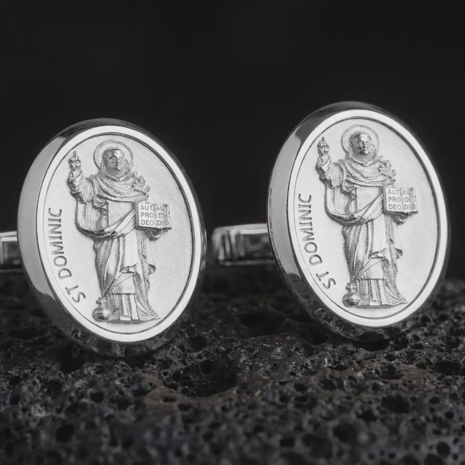Polished silver St Dominic cufflinks are displayed on black volcanic rock in a premium macro studio close-up with detailed Christian devotional artwork.