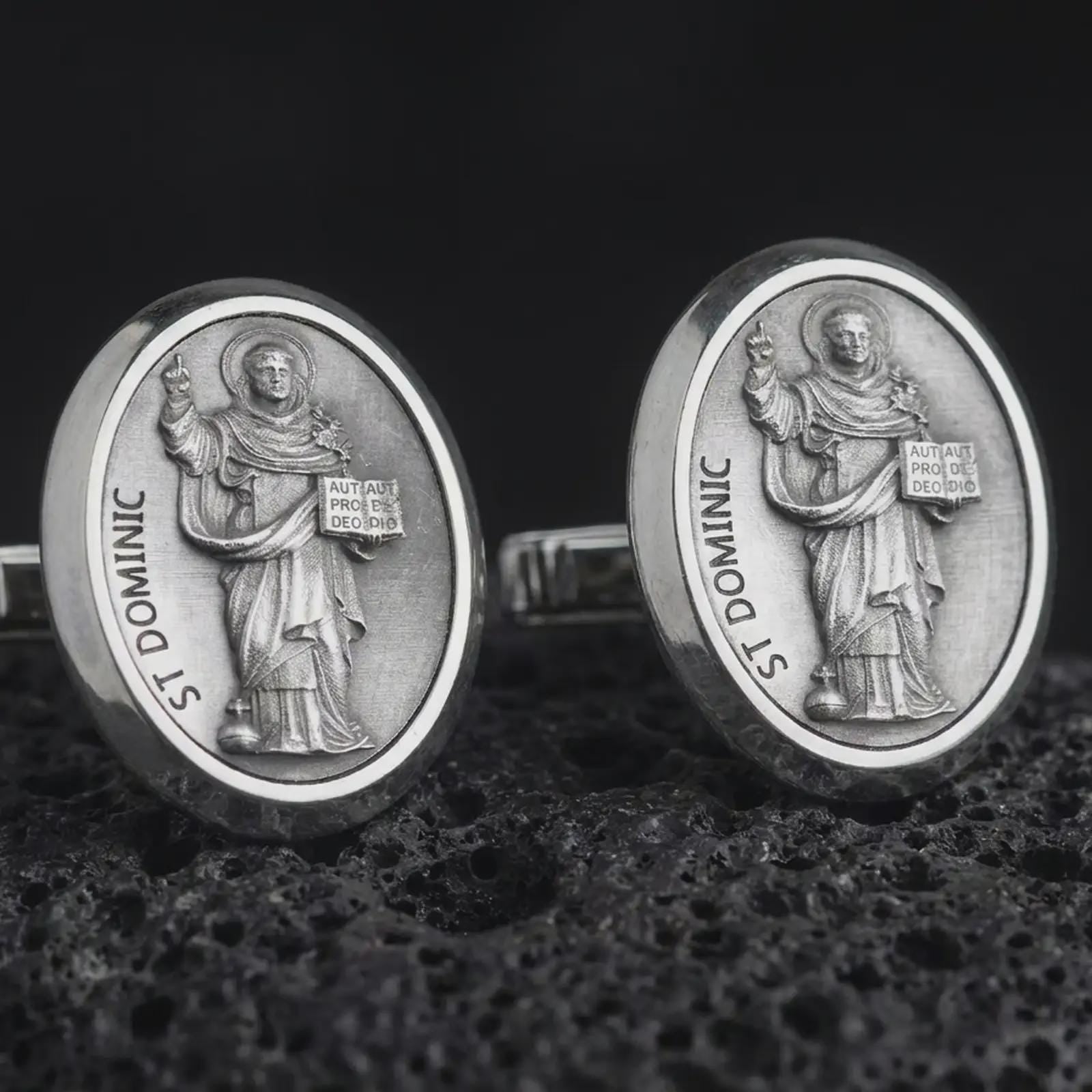 An oxidized St Dominic cufflink pair rests on dark volcanic rock in a macro studio close-up, showing a detailed Catholic saint relief inside oval frames.