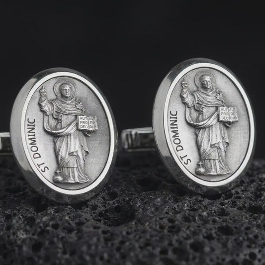 An oxidized St Dominic cufflink pair rests on dark volcanic rock in a macro studio close-up, showing a detailed Catholic saint relief inside oval frames.