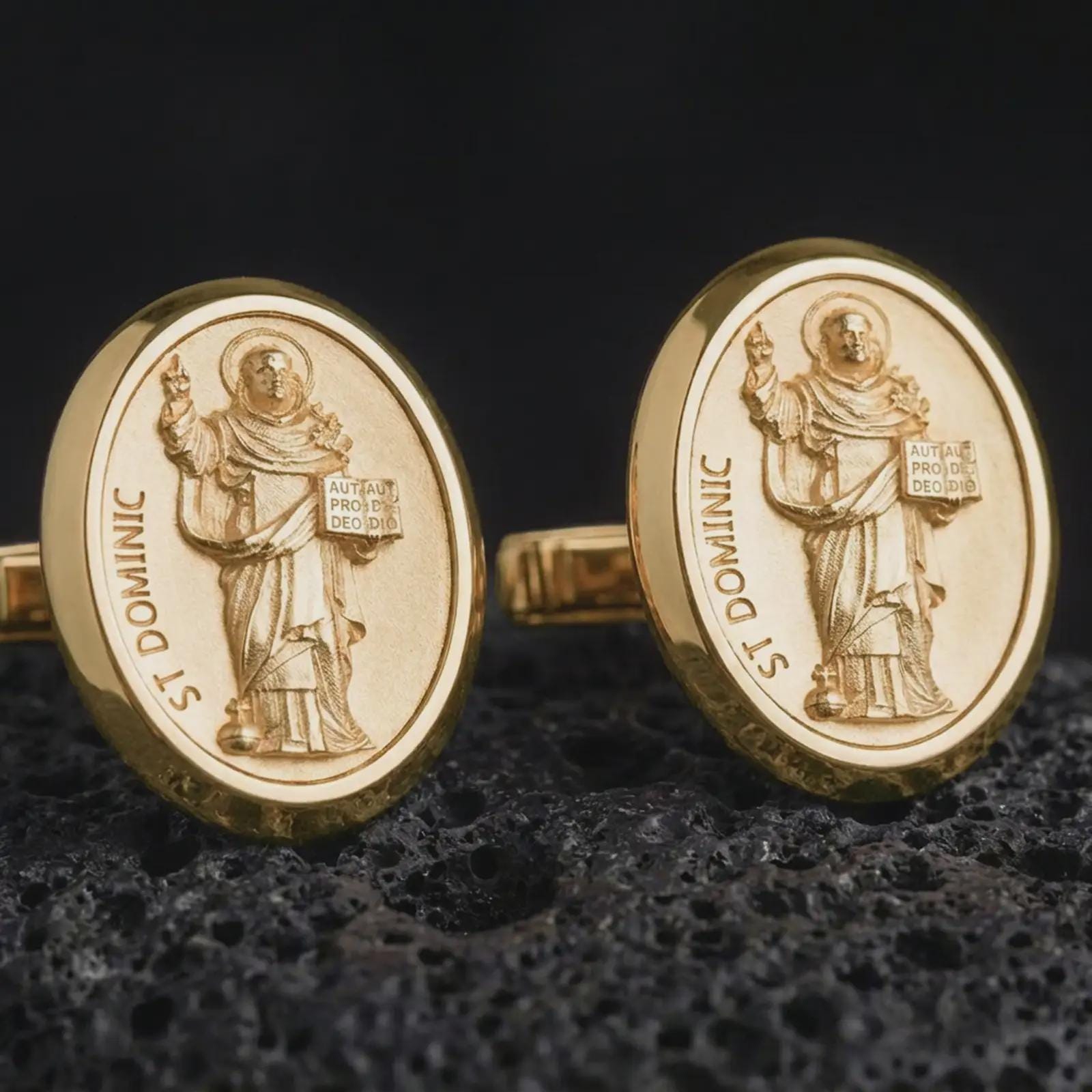 Gold plated St Dominic cufflinks sit on textured volcanic rock in a dramatic studio macro shot, highlighting the sacred saint medallion design.
