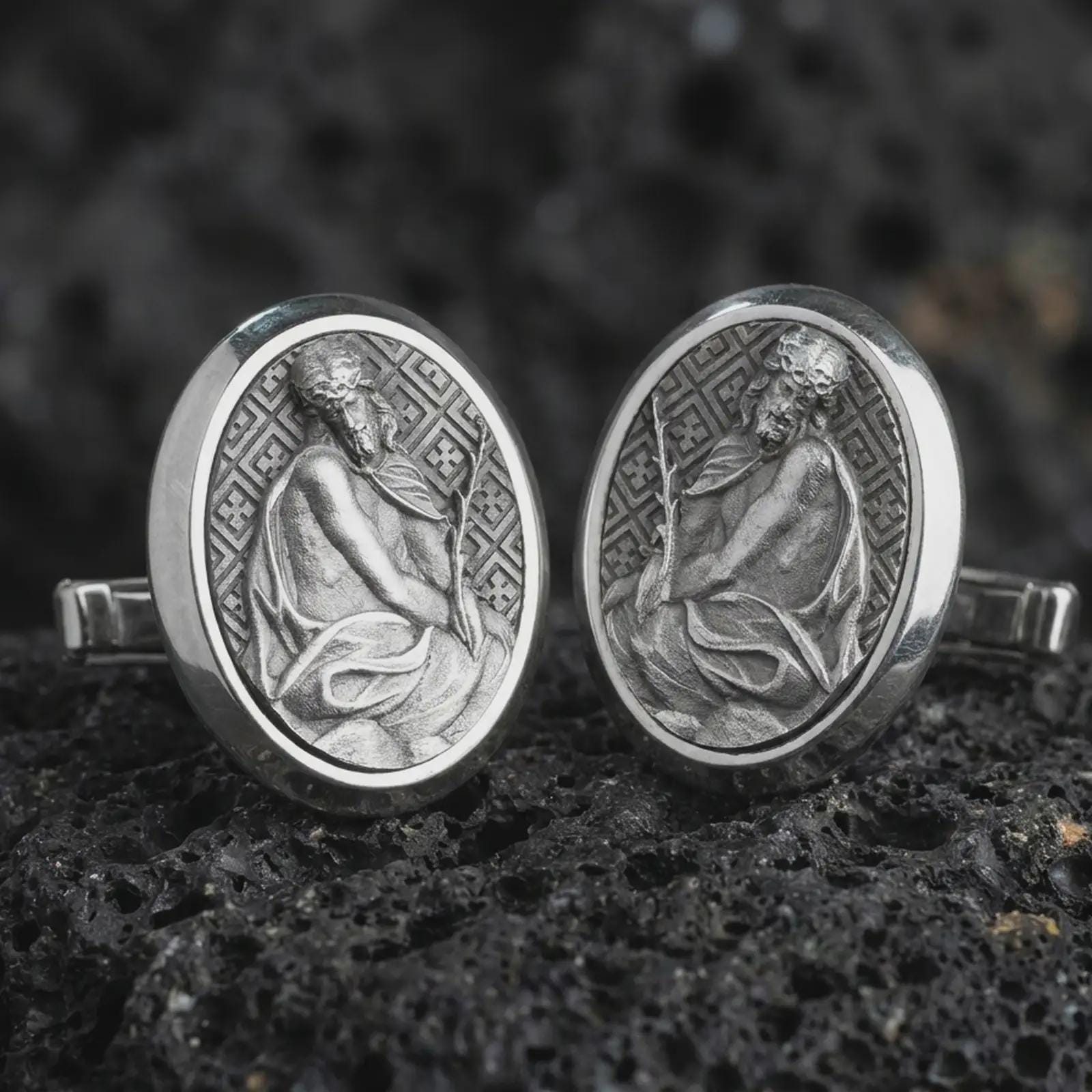 An oxidized Pensive Christ cufflink pair rests on dark volcanic rock in a macro studio close-up, showing a seated Jesus relief inside oval frames.