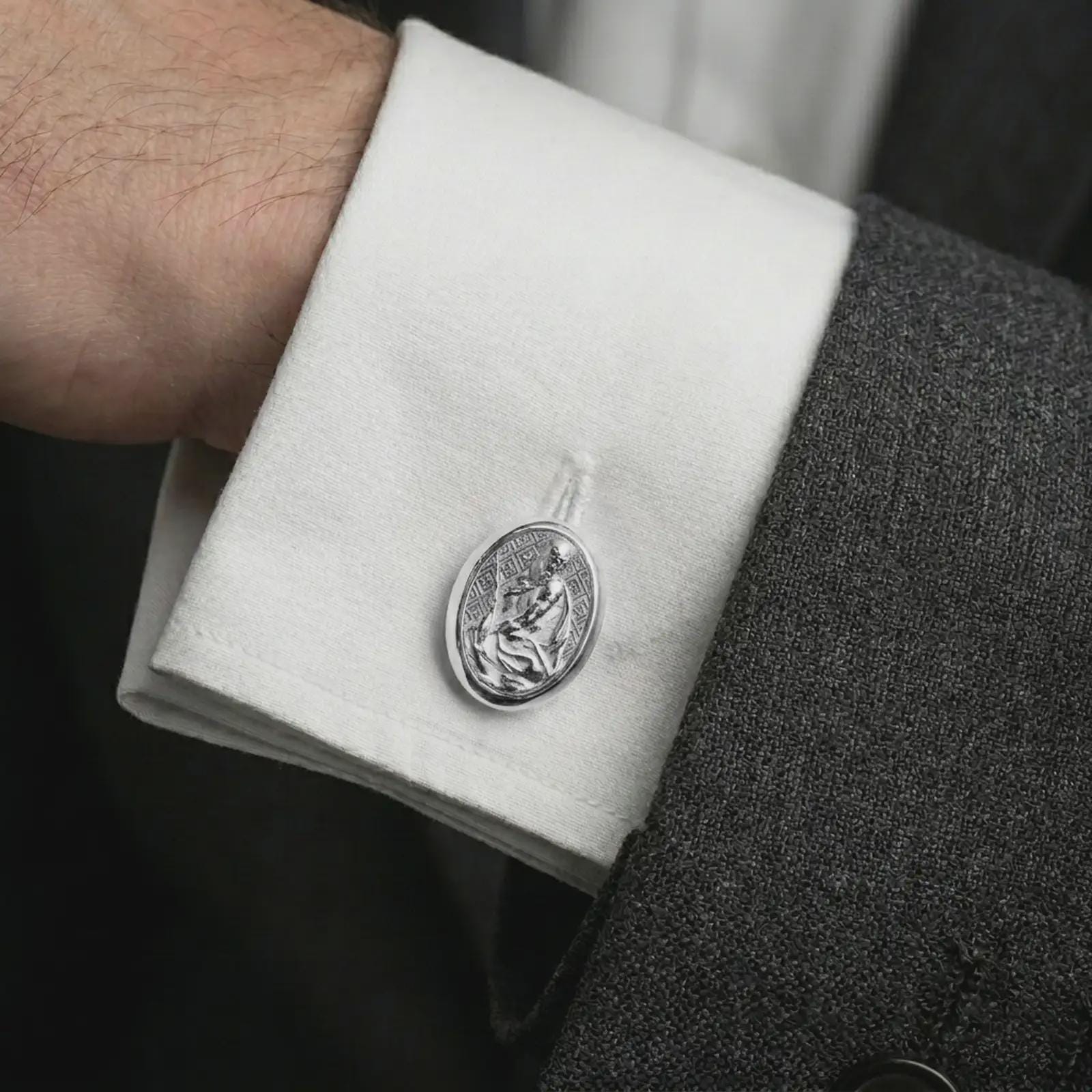 A polished silver Pensive Christ cufflink is worn on a shirt cuff in a premium close-up lifestyle image, showing the seated Jesus relief clearly.