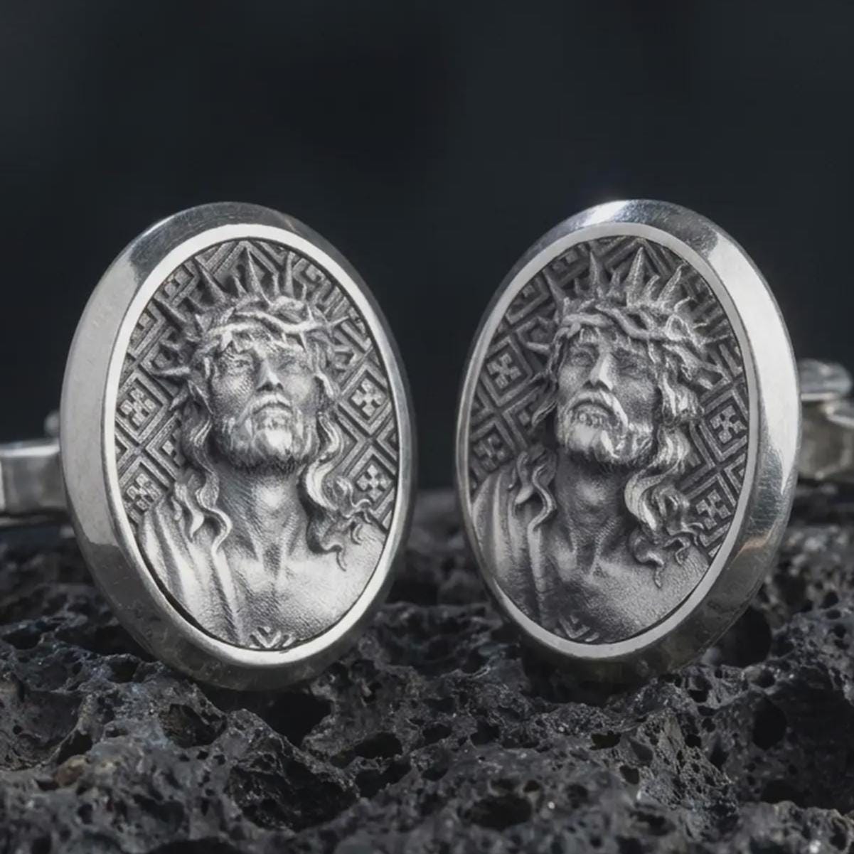 An oxidized Jesus Crown of Thorns cufflink pair rests on dark volcanic rock in a macro studio close-up, showing a detailed Christ portrait inside oval frames.