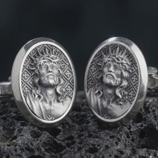 An oxidized Jesus Crown of Thorns cufflink pair rests on dark volcanic rock in a macro studio close-up, showing a detailed Christ portrait inside oval frames.