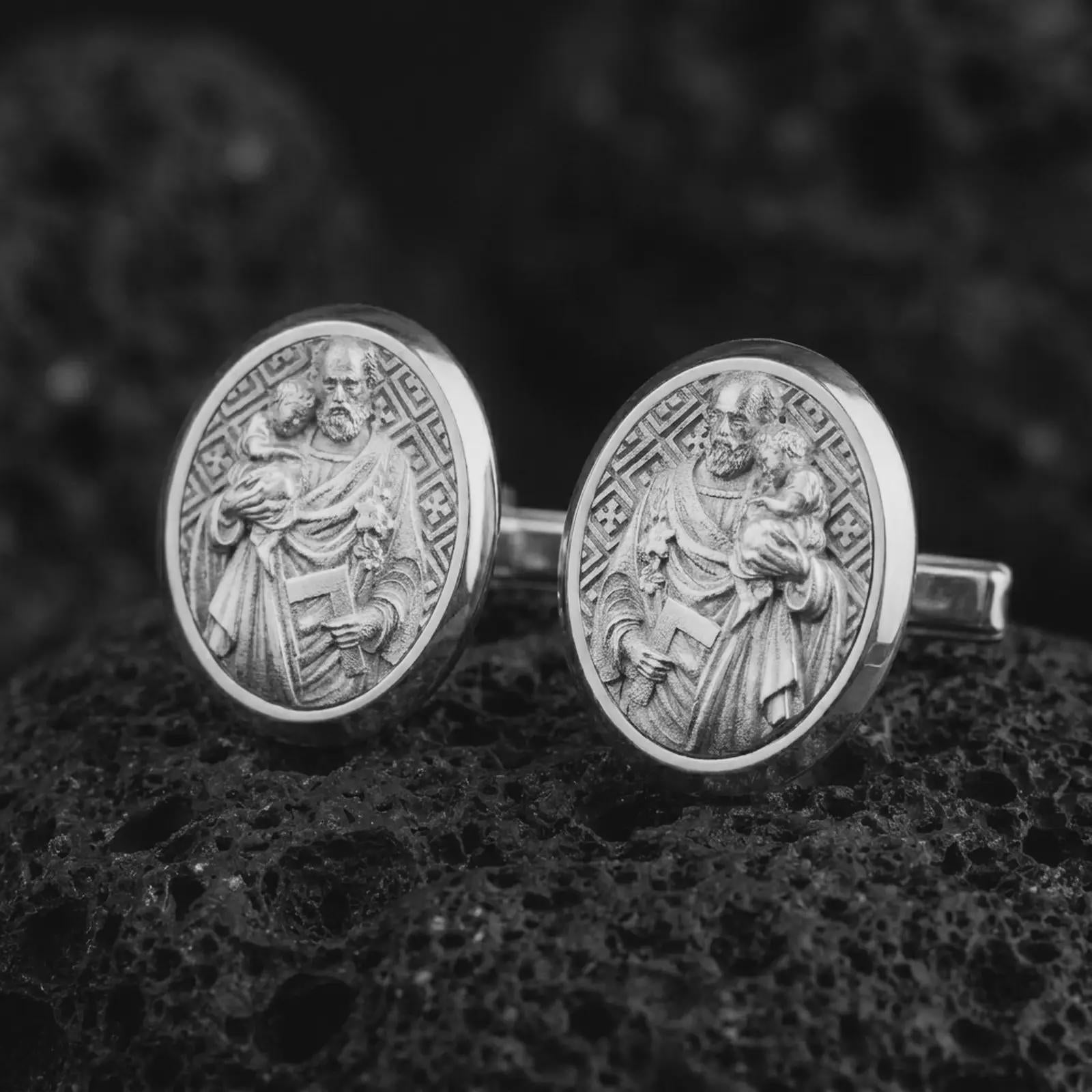 Polished silver Saint Joseph cufflinks resting on textured volcanic rock in a premium macro composition, highlighting the detailed religious relief and oval medallion design.