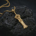 Load image into Gallery viewer, A gold plated demon slayer sword pendant lies on textured volcanic rock in a dramatic studio macro shot, highlighting the hell blade inspired weapon design.
