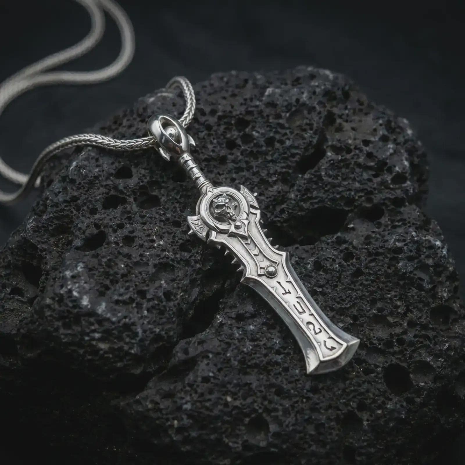 A polished silver demon slayer sword pendant is displayed on black volcanic rock in a premium macro studio close-up with sharp sculpted edges and an ornate guard.