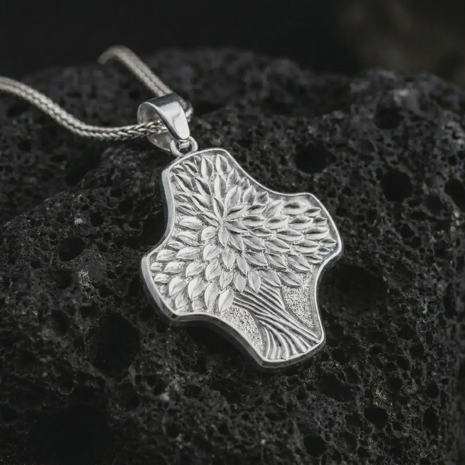 A polished silver Tree of Life cross pendant is displayed on black volcanic rock in a premium macro studio close-up with detailed leaves and flowing branch texture.