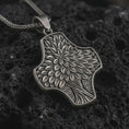Load image into Gallery viewer, An oxidized Tree of Life cross pendant rests on dark volcanic rock in a macro studio close-up, showing layered leaf details and an organic botanical shape.
