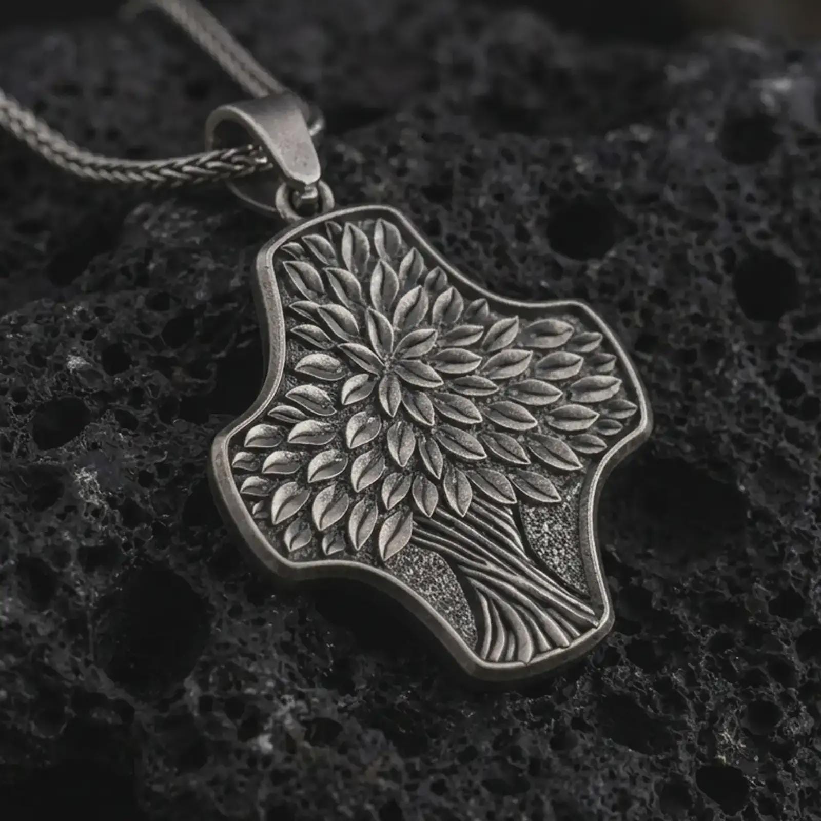 An oxidized Tree of Life cross pendant rests on dark volcanic rock in a macro studio close-up, showing layered leaf details and an organic botanical shape.
