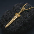 Load image into Gallery viewer, A gold plated fantasy sword pendant lies on textured volcanic rock in a dramatic studio macro shot, highlighting the medieval weapon-inspired design.
